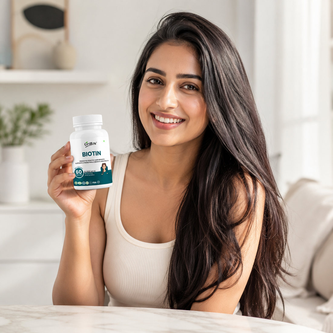 "A smiling woman with long, healthy dark hair sitting in a bright room, holding up a white bottle of Oribite Biotin Capsules."